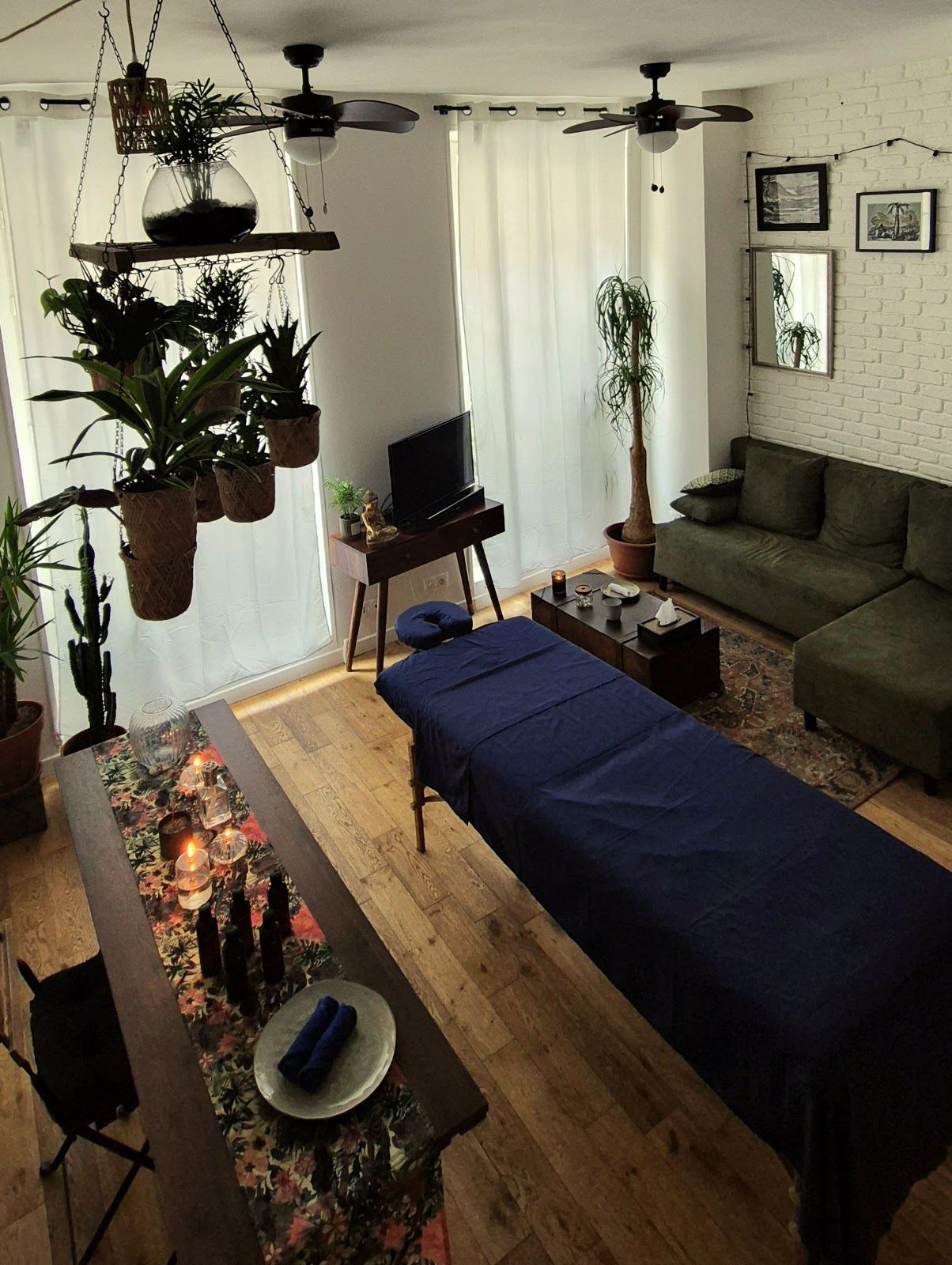 Le même salon en plein jour : table drapée de bleu nuit, parquet clair, plantes suspendues et canapé vert olive